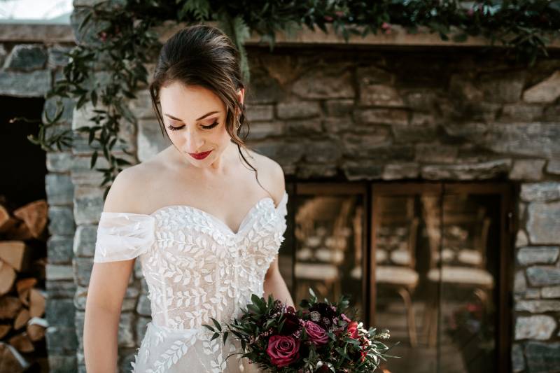 Woman in white lace dress looks down holding dark red bouquet in front of stone fireplace 