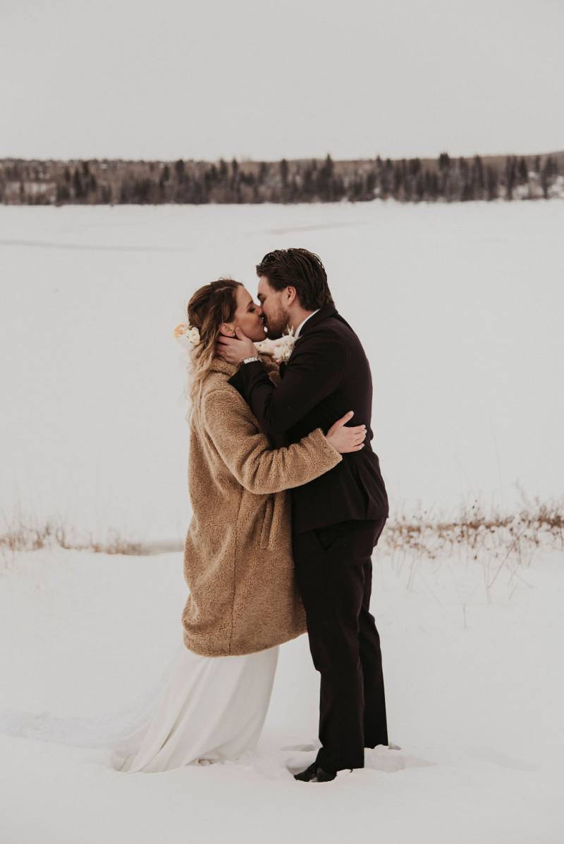 Bride and groom kiss while embracing in snowy field