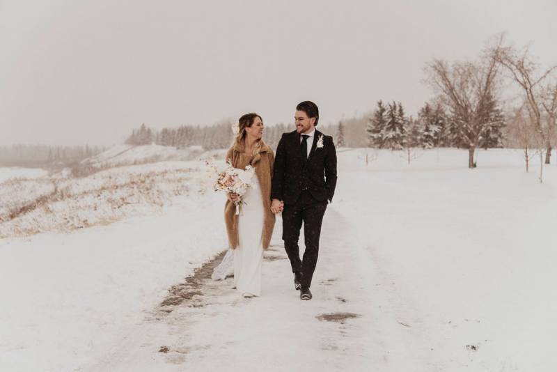 Bride and groom walk down snowy path smiling holding hands