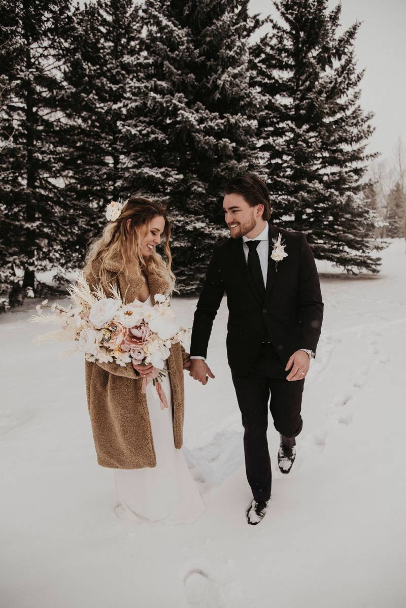 Bride and groom smiling walking down snowy forest path holding bouquet