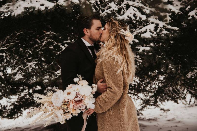 Bride and groom kiss embracing holding bouquet in front of snowy tree 