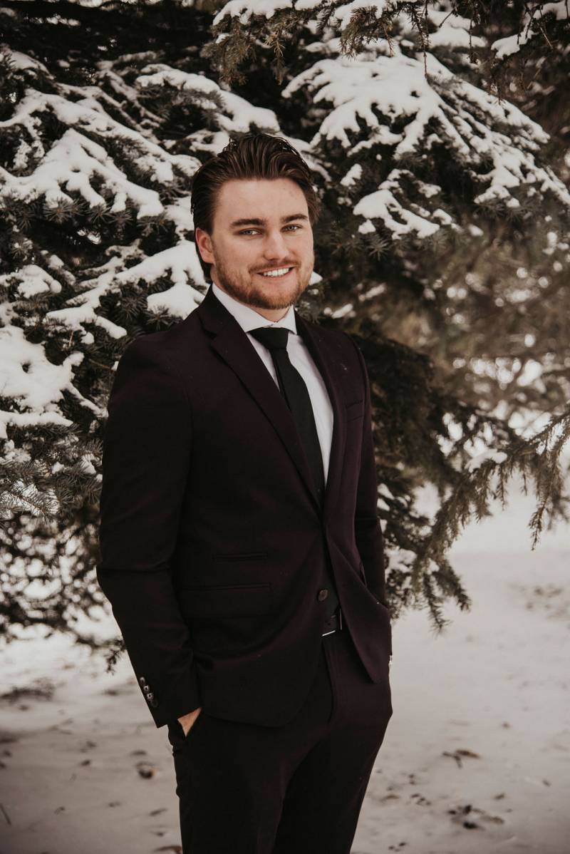 Groom in black suit stands hands in pocket in front of snowy tree