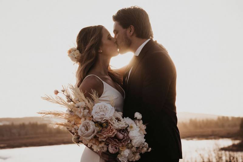 Bride and groom kiss in front of setting sun holding pale pink and white bouquet