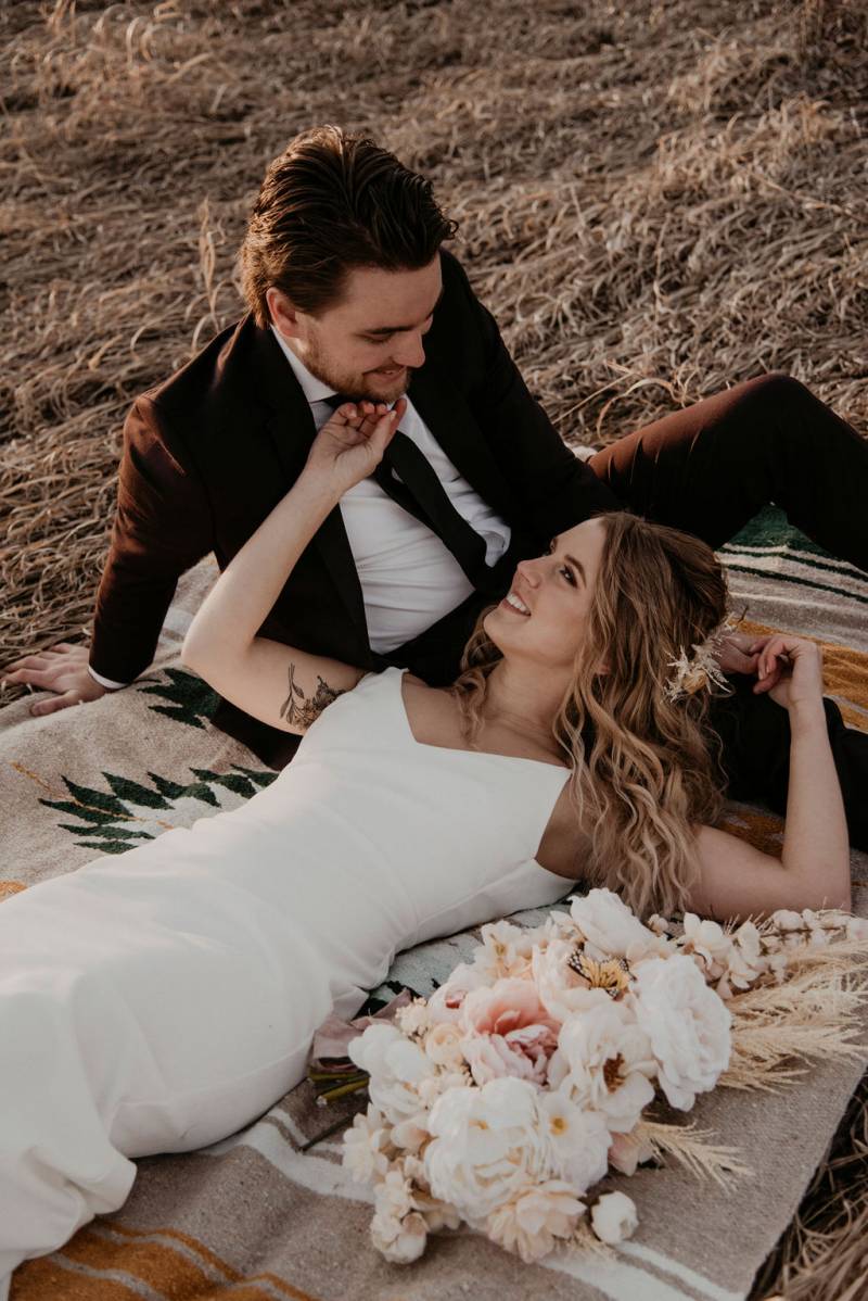 Bride laying in lap of groom in brown grass field on brown tapestry