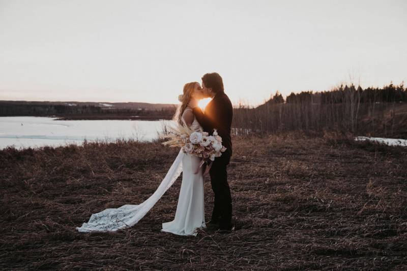 Bride and groom kiss holding bouquet standing in brown grass at dawn 