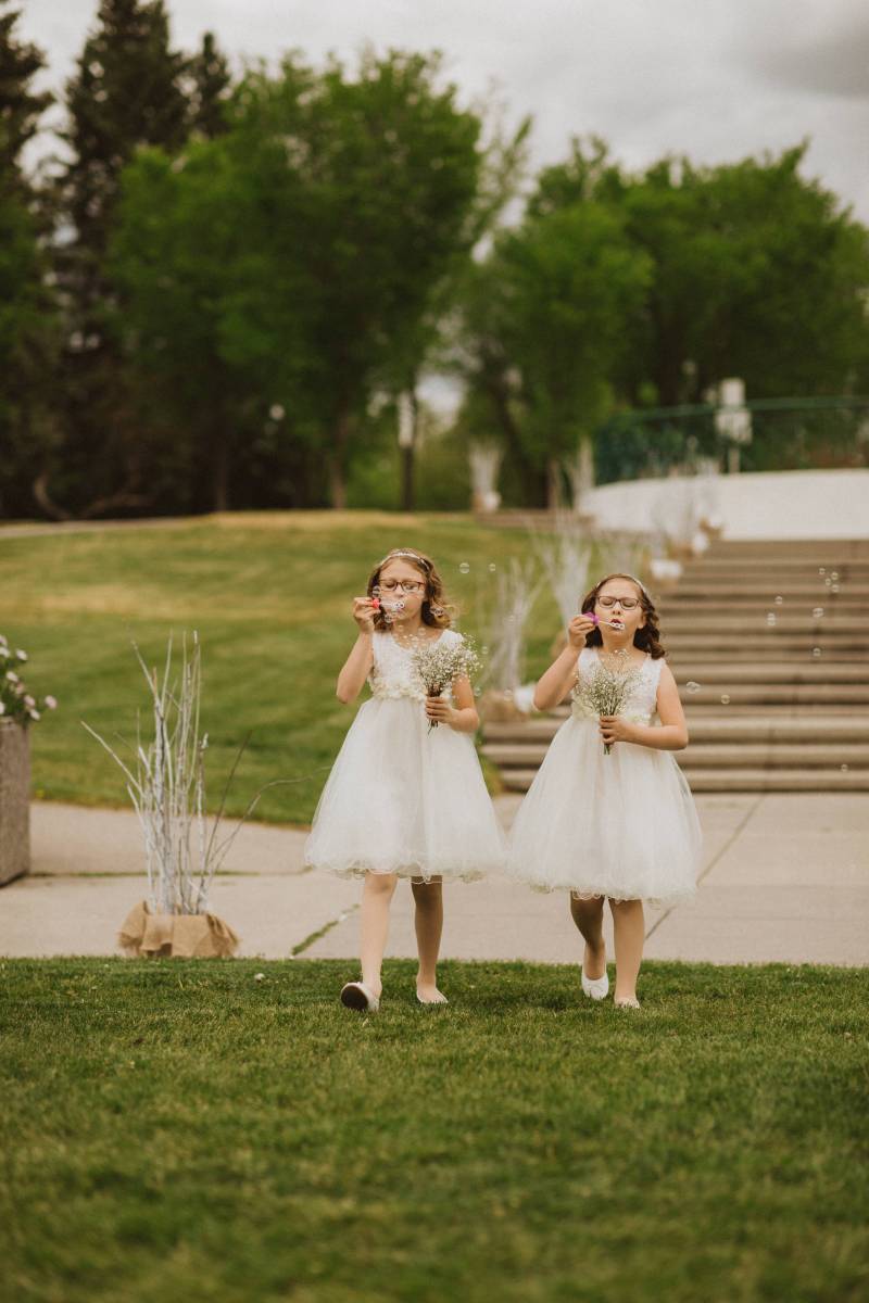 Two flower girls in white dresses holding small bouquets walking blowing bubbles 