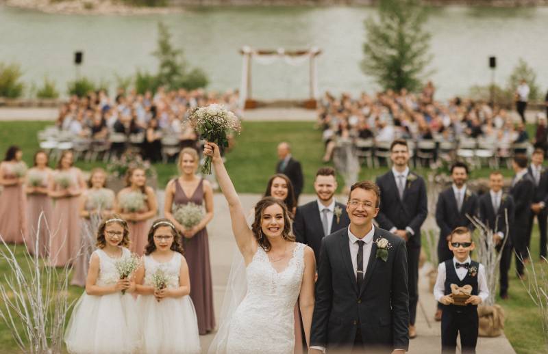 Bride holds up hand with white bouquet while walking in hands with groom guests seated behind 