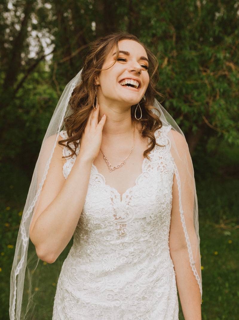 Bride touches neck while smiling in white lace dress and veil 