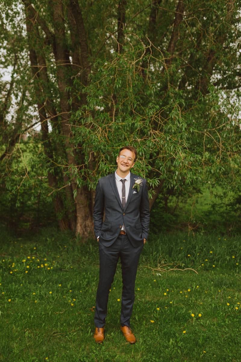 Groom stands hands in pockets smiling in grassy forested area