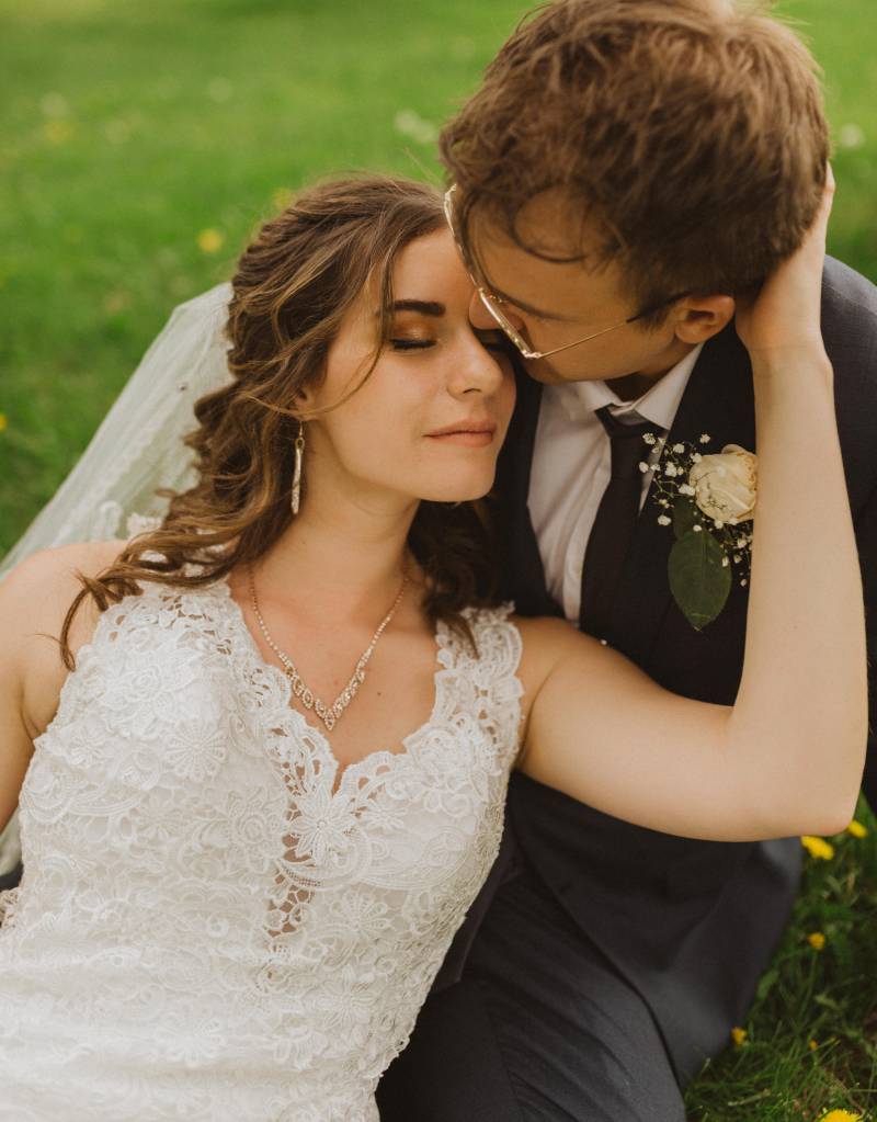 Bride sits in grooms lap touching neck in grassy field 