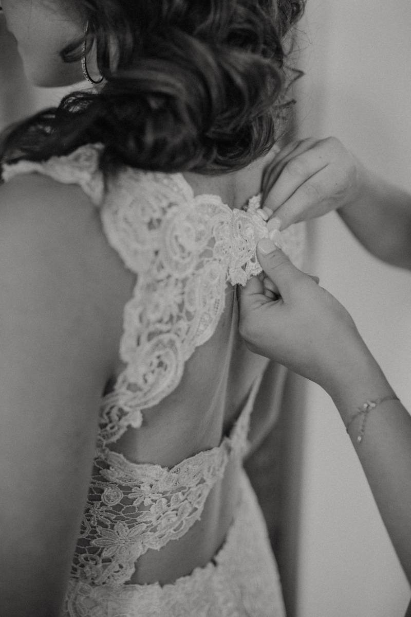 Woman helps bride pin up back of white lace  wedding dress 