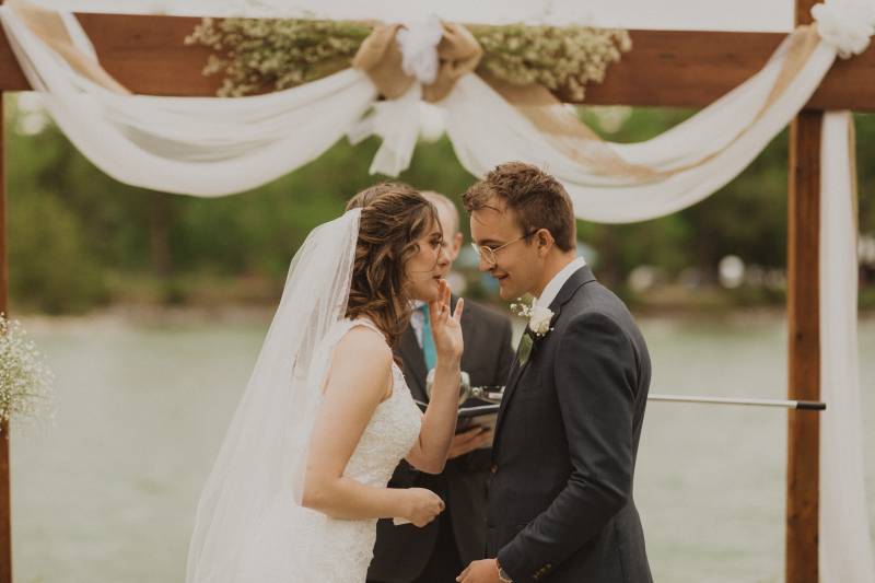 Bride whispers something to groom in front of wedding arch with hanging white fabric