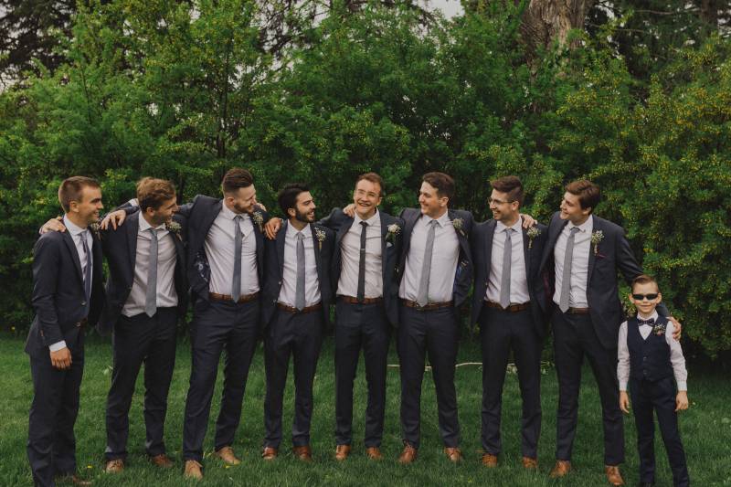 Groom and groomsmen stand arms over shoulders in grassy field 