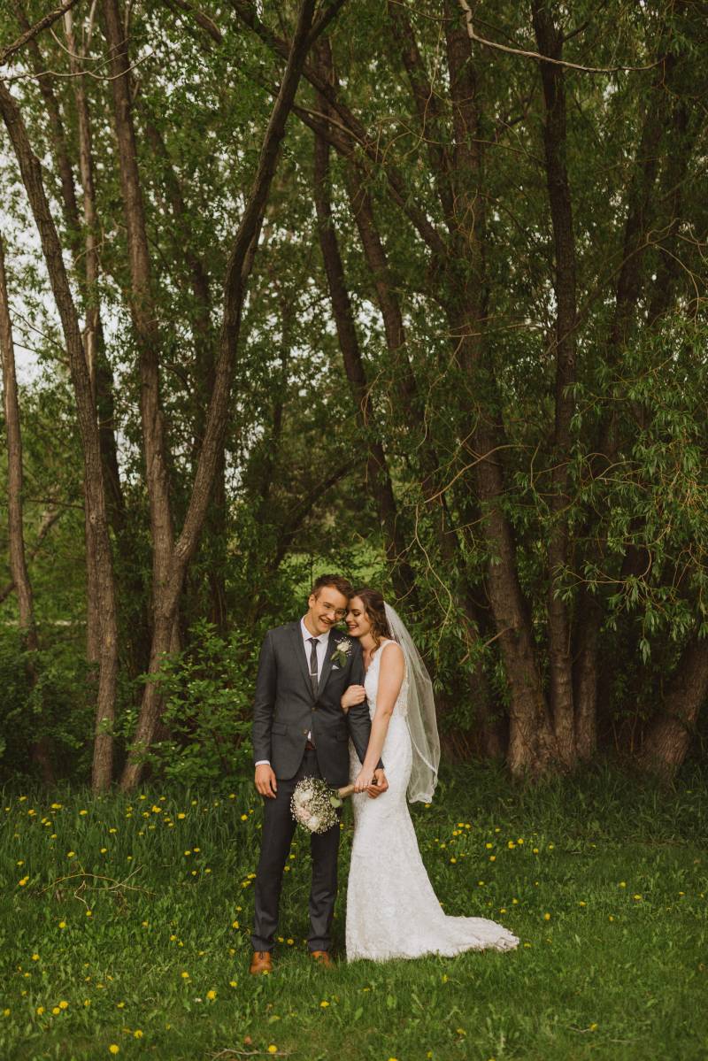 Bride wrapped around grooms arm holding white bouquet in grassy forested area