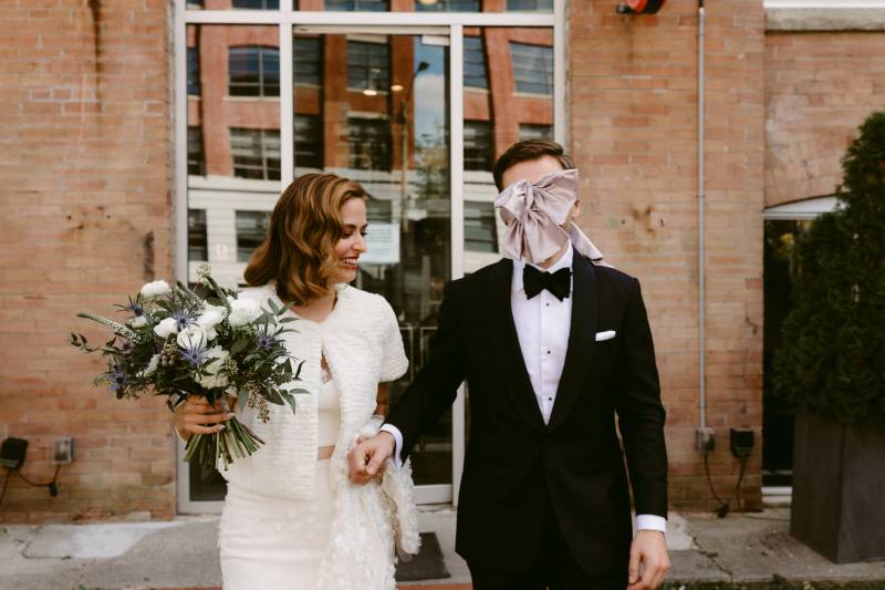 Bride holding bouquet and groom walk outside brick building together while groom wears pastel bow covering eyes 