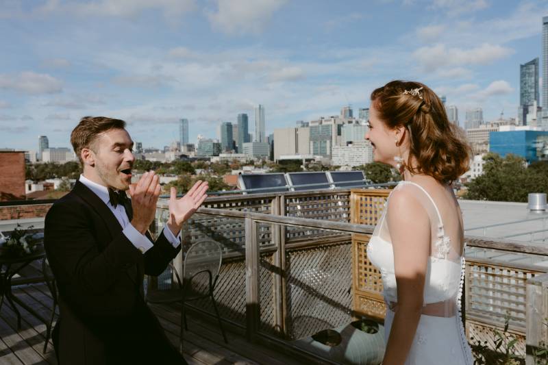 Groom exclaims clapping while looking at bride in white dress on building terrace  
