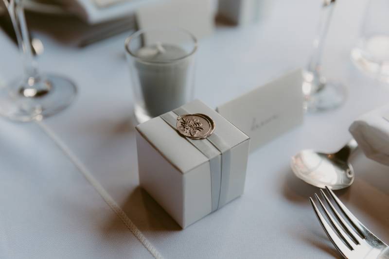 pale grey wedding favor box with bronze wax stamp beside table setting 