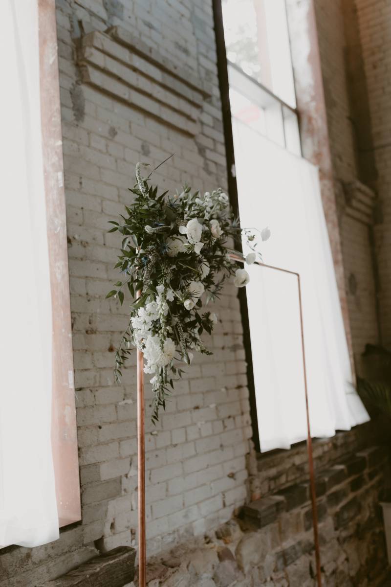 White floral arrangement hanging on copper wedding arch in front of brick wall 