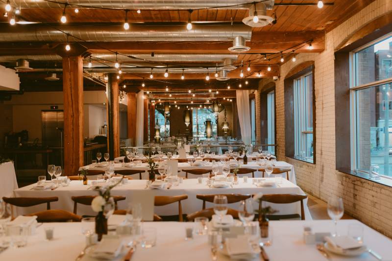 Wedding reception in brick room with fairy lights and large wooden beams over long white tables 