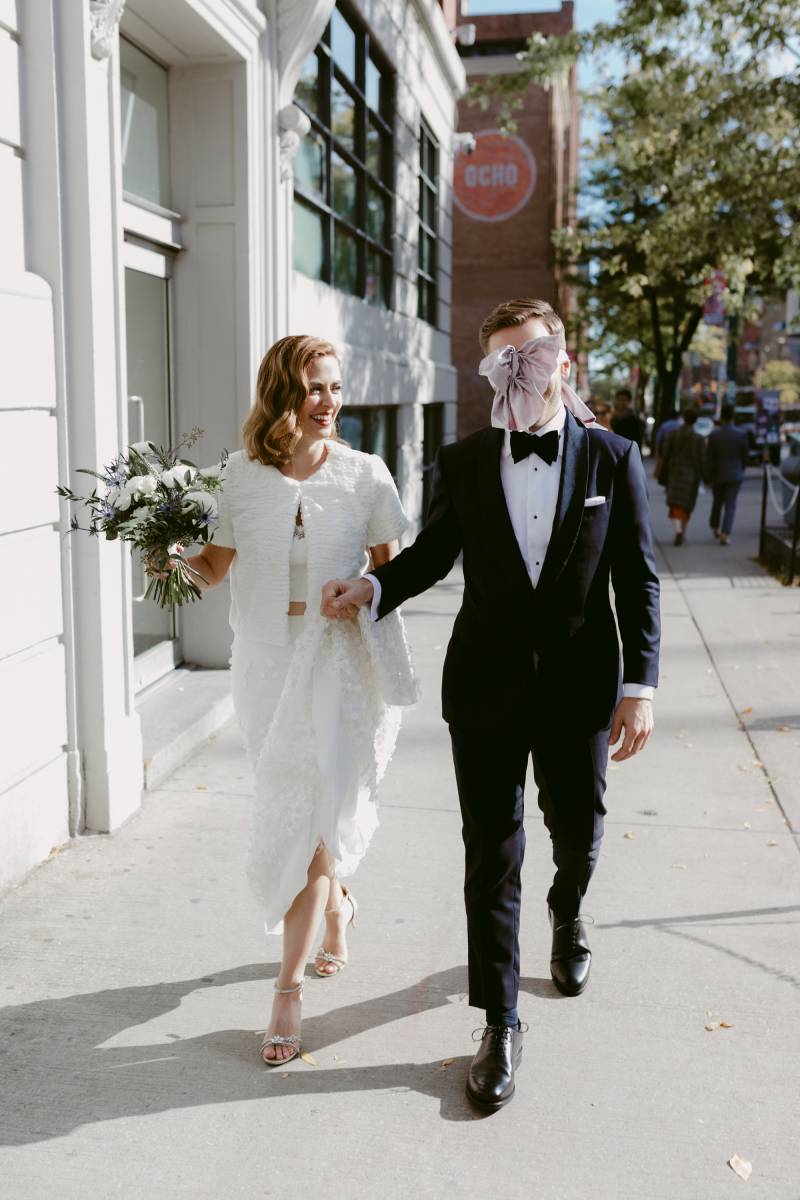 Bride holding bouquet and grooms hand walk on sidewalk together while groom wears pastel bow covering eyes 
