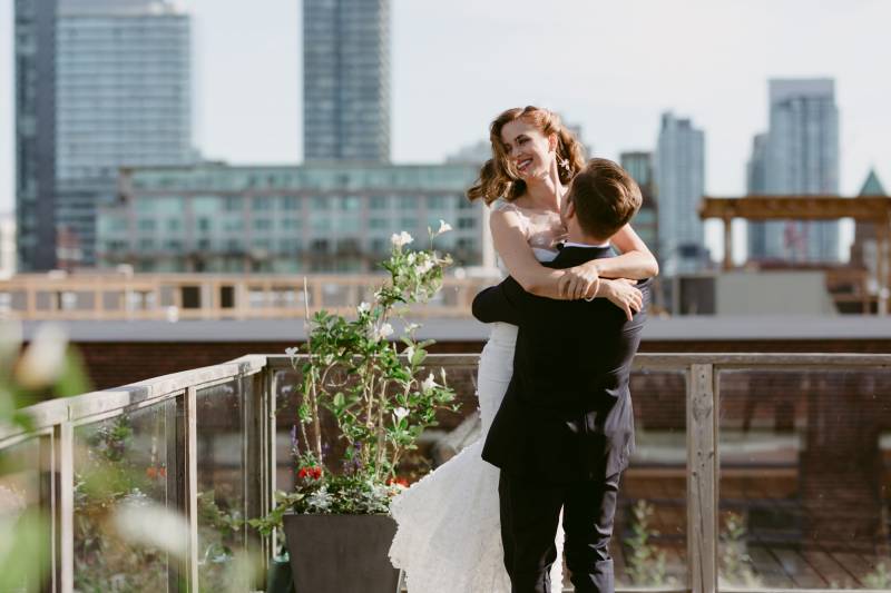 Groom lifts bride in white dress on terrace facing city beside white flower potted plants 