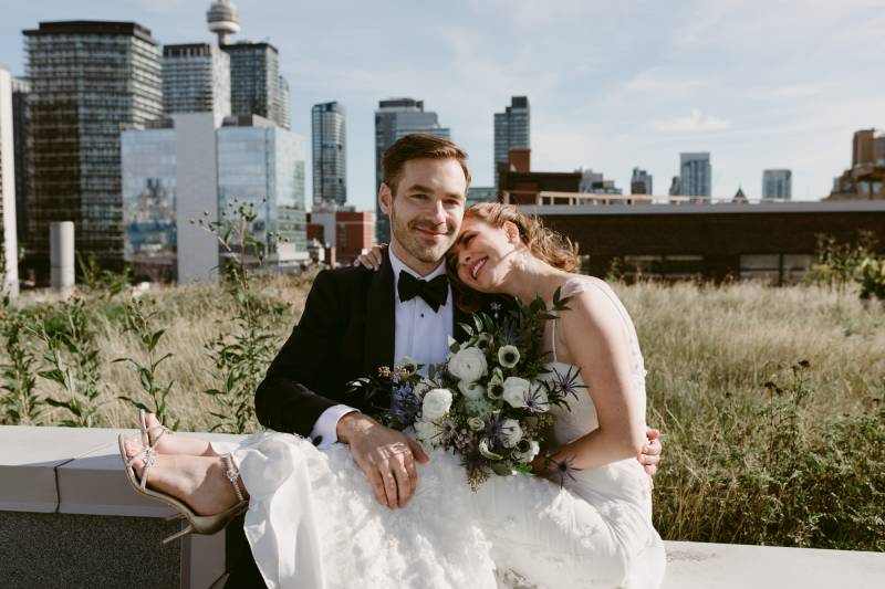 Bride holding white bouquet wearing white dress sitting on grooms lap head on shoulder backing buildings 