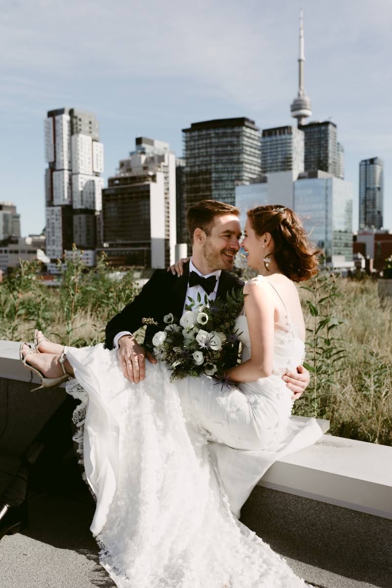 Bride smiling holding white bouquet wearing white dress sitting on grooms lap backing buildings 