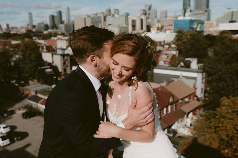 Groom embraces bride kissing cheek while bride in white dress smiles in front of city