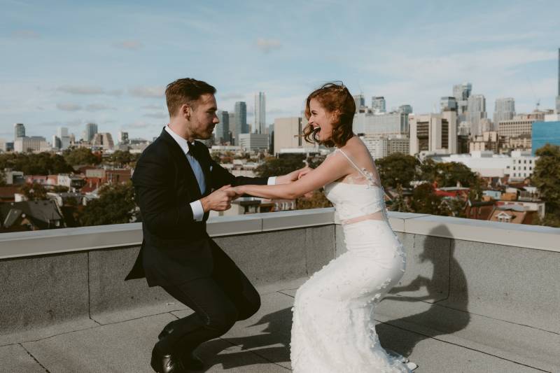 Bride and groom squatting holding hands on roof terrace facing city
