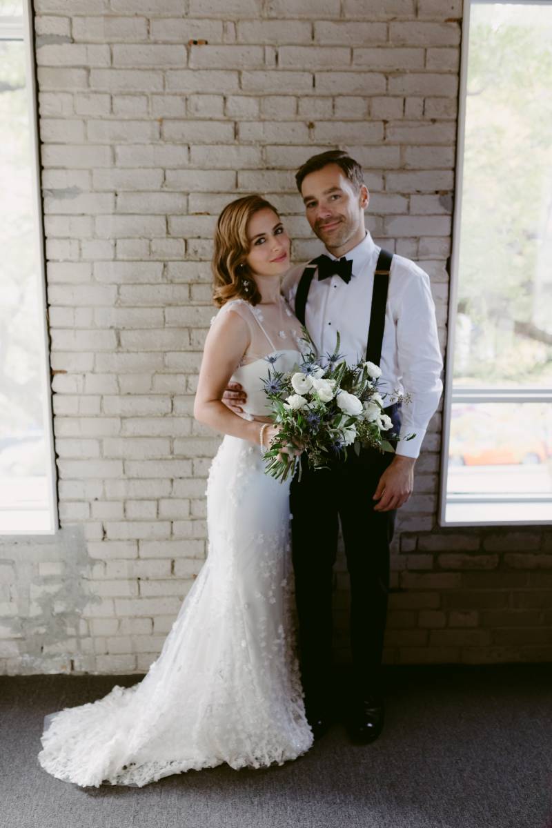 Bride and groom in suspenders stand together on brick wall holding white bouquet 