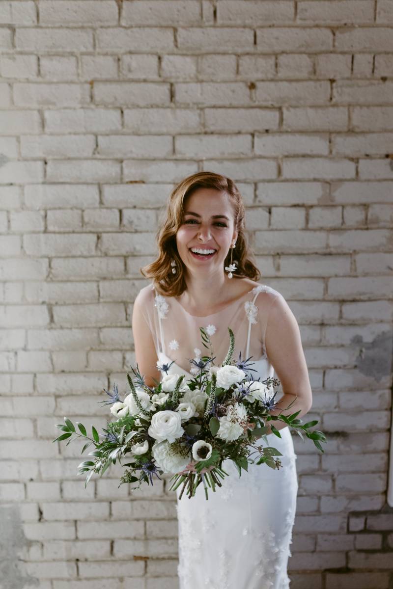 Bride in white dress holding white and lavender bouquet smiling in front of brick wall  