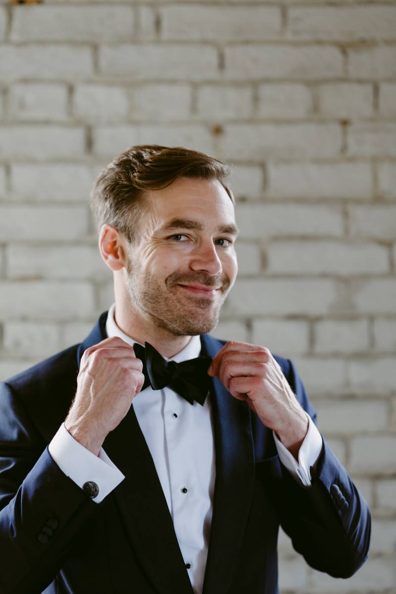 Groom in black and white suit smiling adjusting bowtie in front of grey brick wall