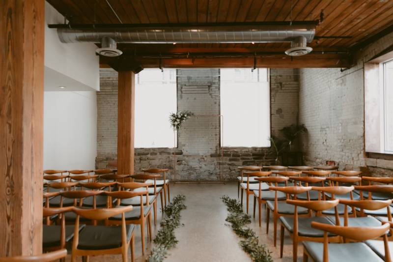 Small brick room with light brown wooden chairs facing copper wedding arch with white floral accents