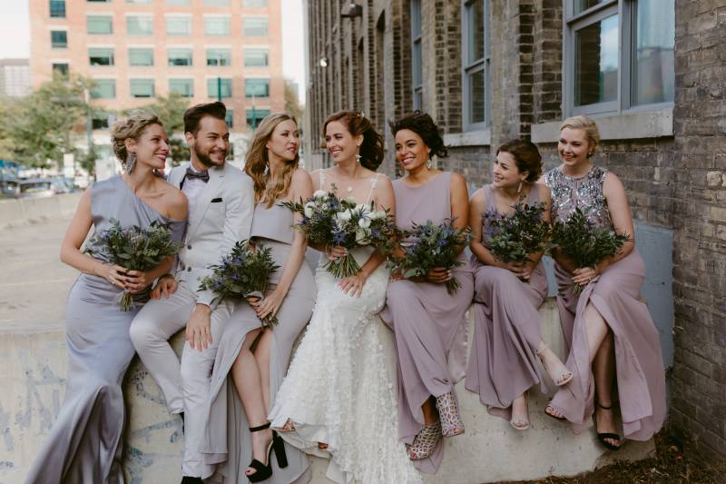 Bride and bridesmaids sit together in line holding lavender and white bouquets outside brick building 