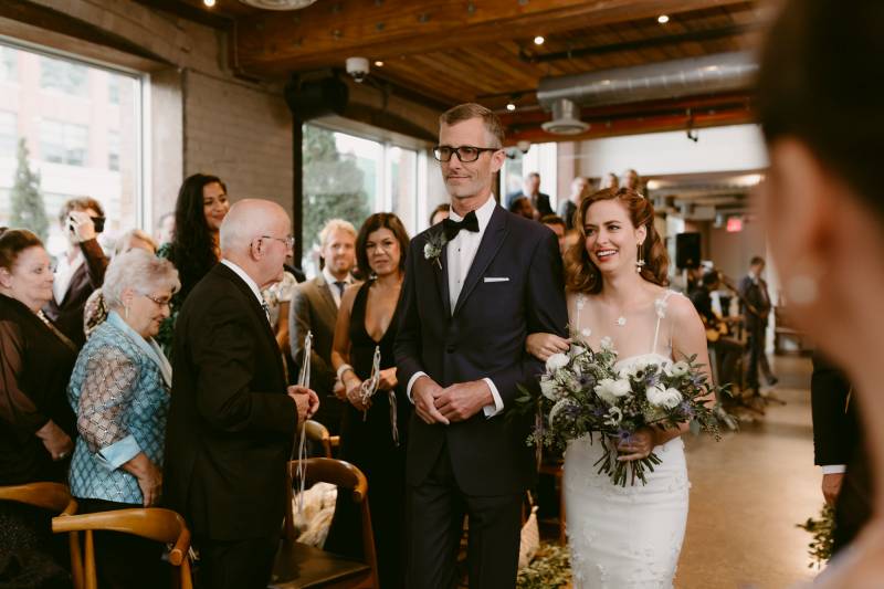 Bride holding white bouquet walking down aisle in arms with man in suit smiling while guests watch 