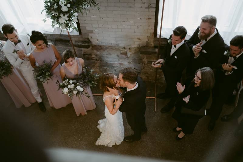 Bride and groom kiss while embracing in white lace dress with white floral accents as guests applaud 