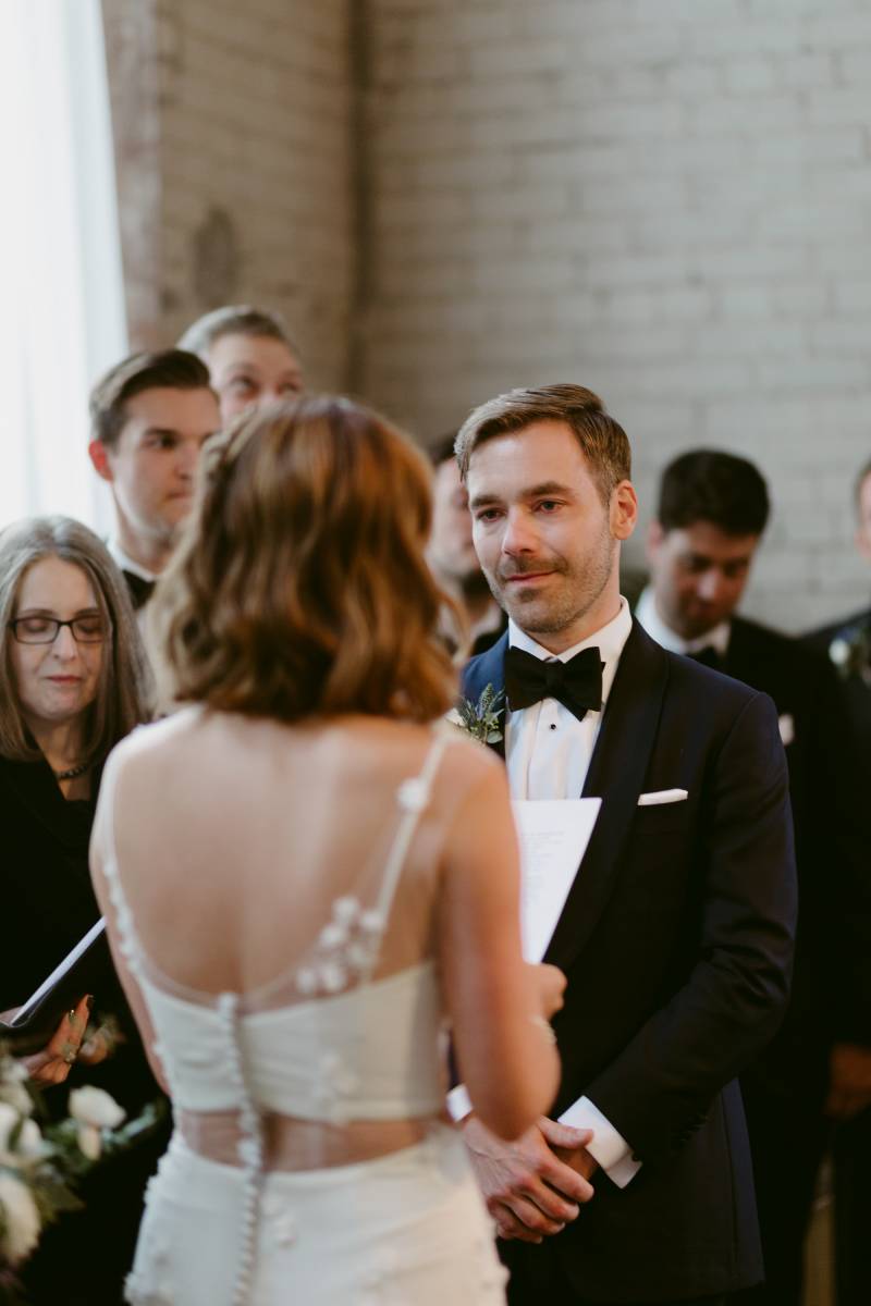 Bride and groom stand facing in white lace dress with white floral accents reading vows 