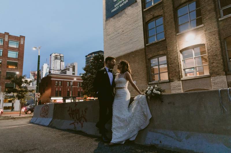 Bride and groom stand together against concrete barrier outside 