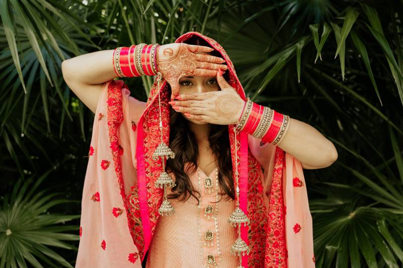 Woman with henna holds  hands in front of face revealing eyes in traditional red and peach dress 