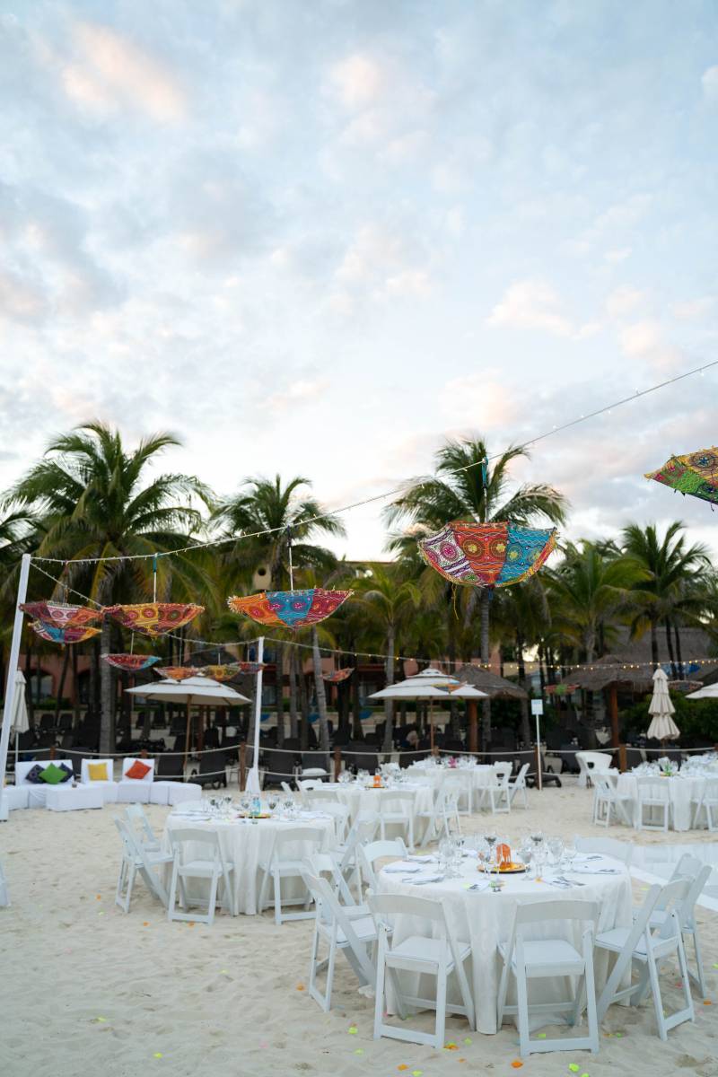 White tables set with white chairs on beach under hanging umbrellas  
