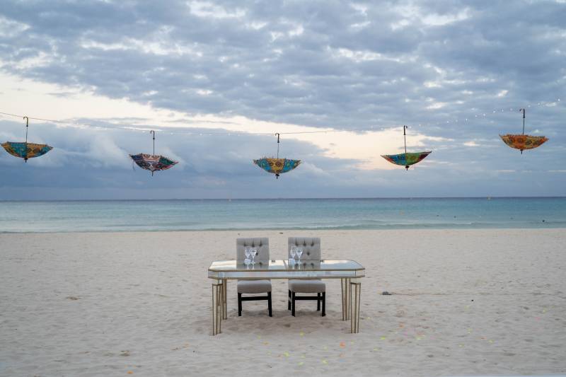 Clear glass sweetheart table  on beach with hanging multicolored umbrellas on fairy light line