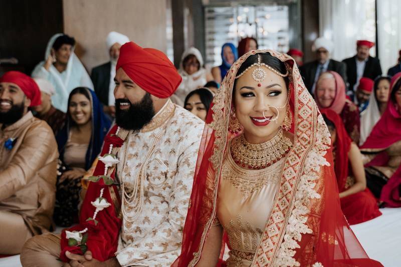 Man and woman sit in red traditional wedding attire smiling with guests sitting behind