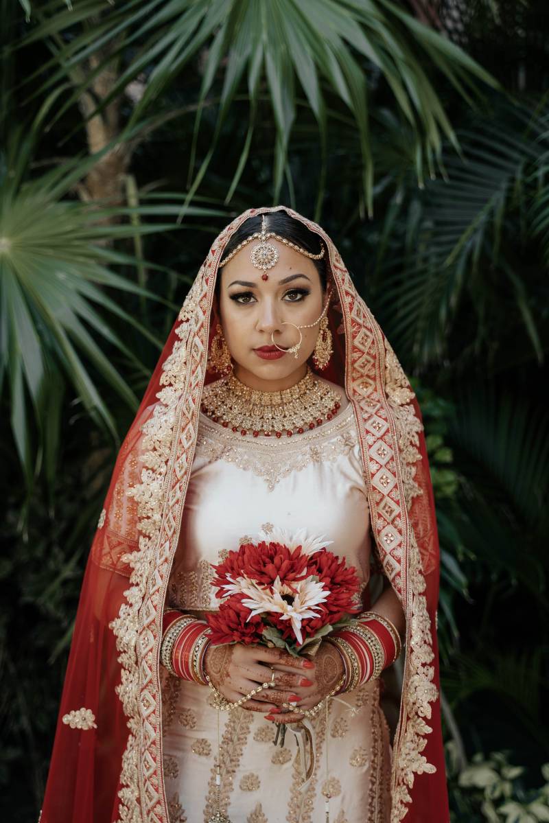 Woman stands in crimson and cream traditional wedding dress holding white and red bouquet in front 