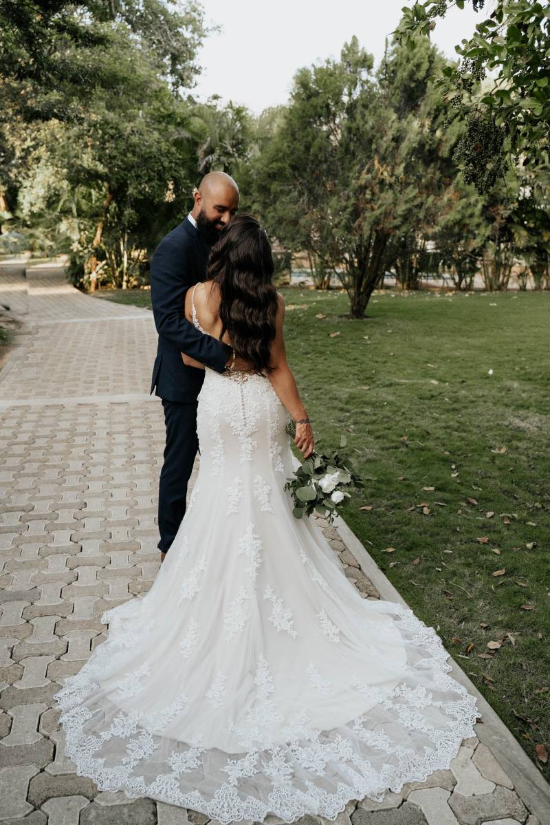 Groom embraces bride wearing large white lace dress holding white bouquet on park sidewalk 