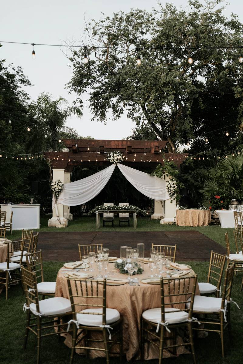 Outdoor wedding reception with hanging lights and circle tables surrounding wood patio facing head table under hanging white fabric awning 