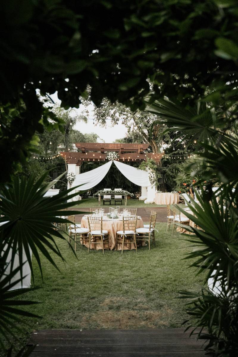 Outdoor wedding reception with hanging lights and circle tables surrounding wood patio facing head table under hanging white fabric awning 