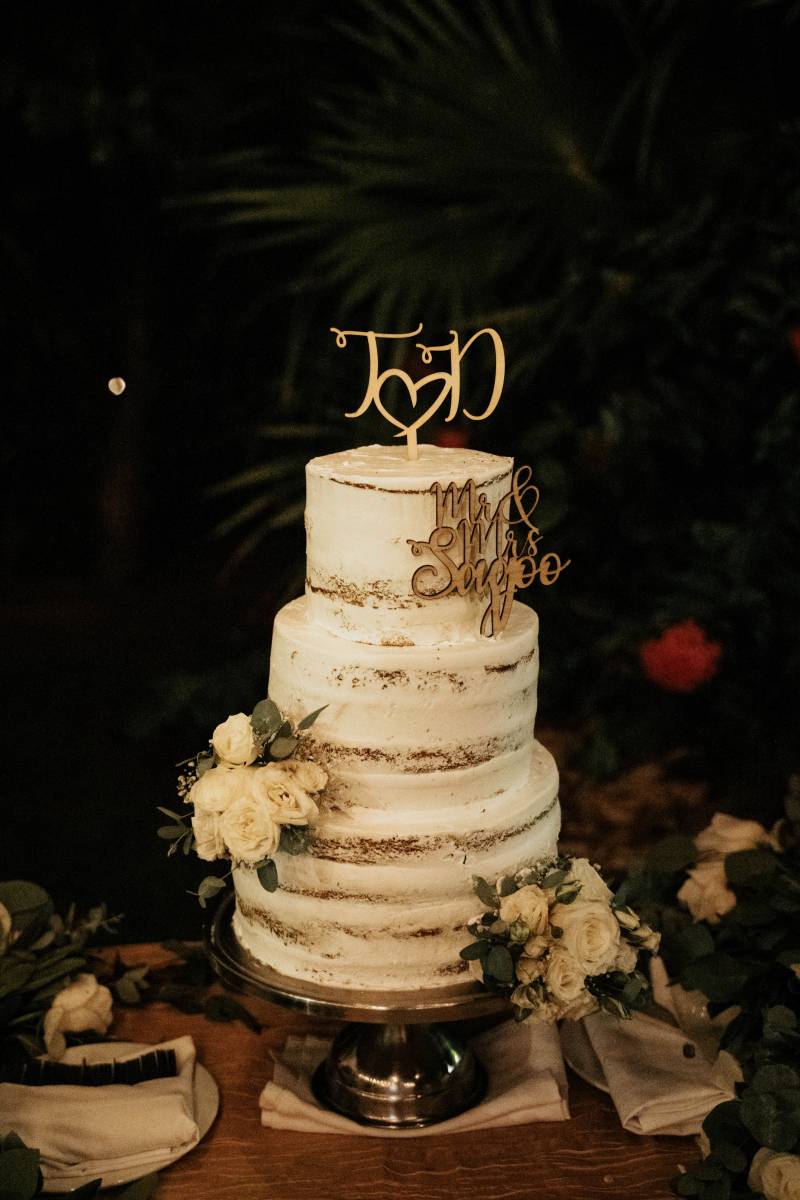 Naked white wedding cake with white floral accents and gold initials on top 