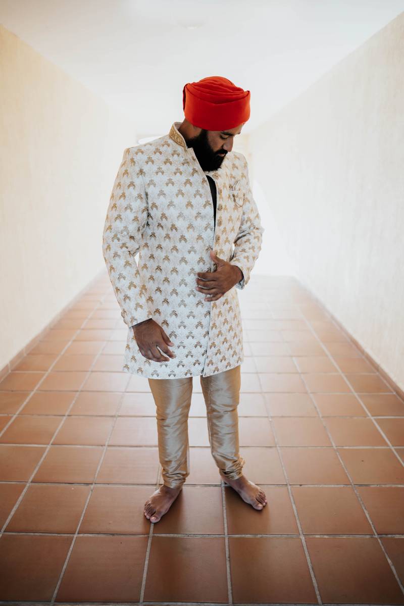 Man stands in red head wrap and traditional white and gold suit and pants 