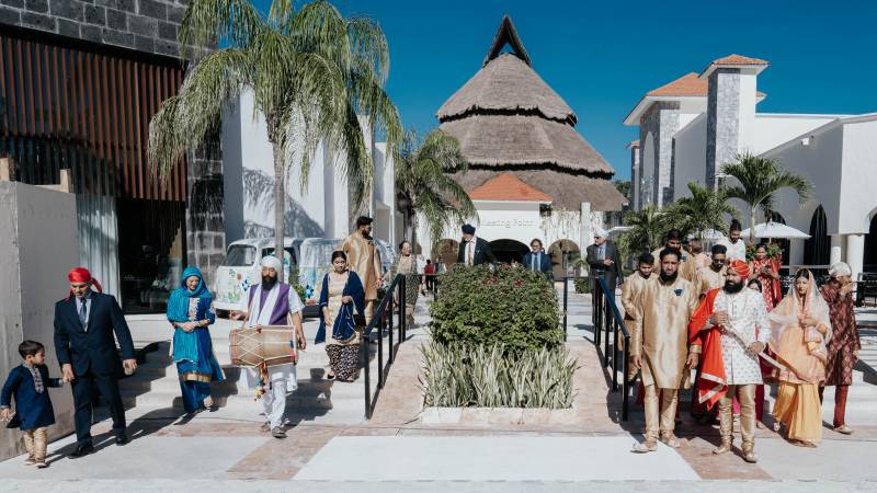 Bride and groom walk amongst guests from building with tapered brown roof and palmtrees