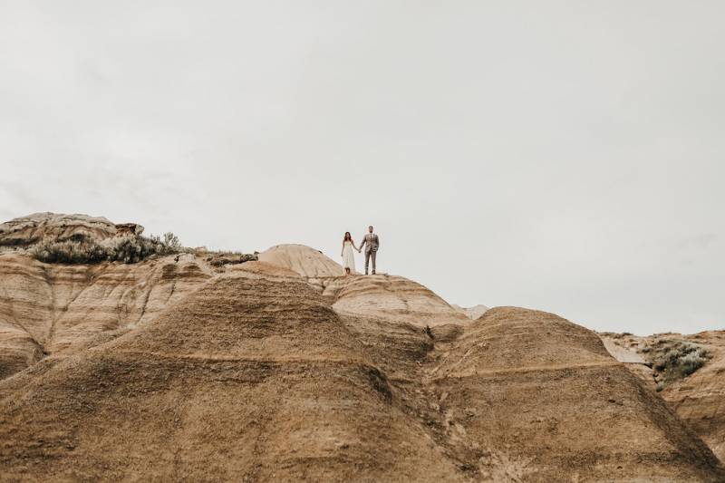 Man and woman stand holding hands at top of large dirt hill 